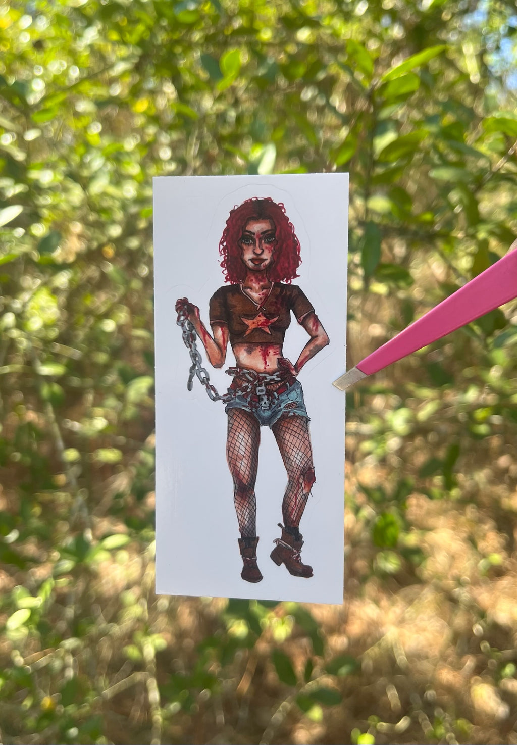 Sticker of a woman zombie fighter with red hair and chains held by a pink pen against a green leafy background